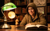 Une personne en sweat à capuche sourit devant un échiquier et un livre dans une bibliothèque au style classique.
