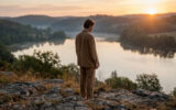 Jeune homme contemplatif face à un lac brumeux au lever du soleil, entouré de collines boisées. Scène paisible.