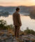 Jeune homme contemplatif face à un lac brumeux au lever du soleil, entouré de collines boisées. Scène paisible.