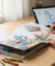 Personne dessinant sur un livre pour enfants ouvert avec un stylet, une tablette affiche un croquis numérique. Matériel artistique sur un bureau.