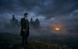 Cillian Murphy, costume et manteau sombres, debout sur une colline herbeuse, contemplant des ruines dans la brume sous un ciel nuageux, avec un feu lointain.