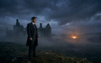 Cillian Murphy, costume et manteau sombres, debout sur une colline herbeuse, contemplant des ruines dans la brume sous un ciel nuageux, avec un feu lointain.