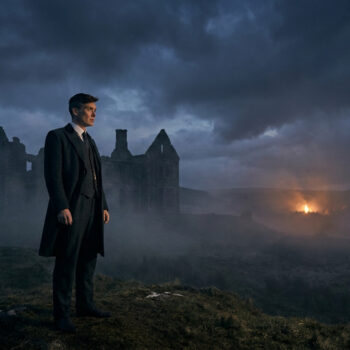 Cillian Murphy, costume et manteau sombres, debout sur une colline herbeuse, contemplant des ruines dans la brume sous un ciel nuageux, avec un feu lointain.