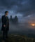 Cillian Murphy, costume et manteau sombres, debout sur une colline herbeuse, contemplant des ruines dans la brume sous un ciel nuageux, avec un feu lointain.