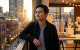 Jeune homme souriant debout sur une terrasse au coucher du soleil avec une vue sur la ville en arrière-plan.