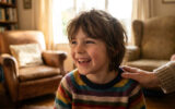 Un enfant aux cheveux longs, vêtu d'un pull rayé, rit joyeusement. Une main adulte le réconforte sur l'épaule dans un salon lumineux.