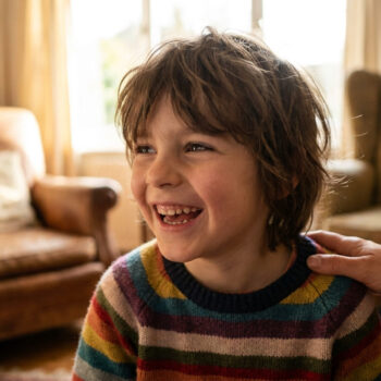 Un enfant aux cheveux longs, vêtu d'un pull rayé, rit joyeusement. Une main adulte le réconforte sur l'épaule dans un salon lumineux.