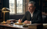 Roger Peyrefitte écrit à un bureau en bois ancien, entouré de livres. Il tient une plume, l'air pensif, sous une lampe de bronze.