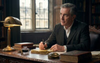 Roger Peyrefitte écrit à un bureau en bois ancien, entouré de livres. Il tient une plume, l'air pensif, sous une lampe de bronze.