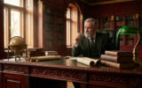 Homme âgé à barbe grise, costume tweed, assis à un grand bureau en bois sculpté dans une bibliothèque, stylo à la main, pensif.