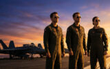 Trois pilotes (deux hommes, une femme) en combinaison de vol et lunettes de soleil sur un porte-avions au coucher du soleil, avec des jets en fond.