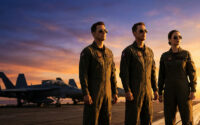 Trois pilotes (deux hommes, une femme) en combinaison de vol et lunettes de soleil sur un porte-avions au coucher du soleil, avec des jets en fond.