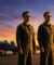 Trois pilotes (deux hommes, une femme) en combinaison de vol et lunettes de soleil sur un porte-avions au coucher du soleil, avec des jets en fond.