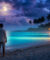 Un homme debout sur une plage tropicale au clair de lune, regardant l'océan la nuit sous un ciel étoilé et nuageux.