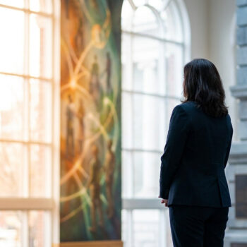 Une femme en tailleur sombre vue de dos, contemplant une fresque murale lumineuse dans un hall d'entrée.