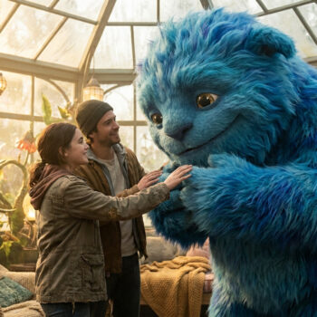 Ryan Reynolds et une jeune fille sourient face à une grande créature poilue bleue dans une serre lumineuse.