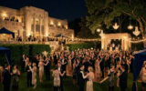 Une foule d'invités en tenues des années 20 danse et socialise lors d'une fête nocturne somptueuse devant un grand manoir.
