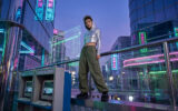 Une personne vêtue d'un sweat chromé debout sur une rambarde dans une ville cyberpunk avec des néons et des ordinateurs.