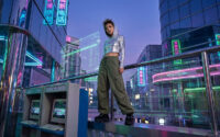 Une personne vêtue d'un sweat chromé debout sur une rambarde dans une ville cyberpunk avec des néons et des ordinateurs.