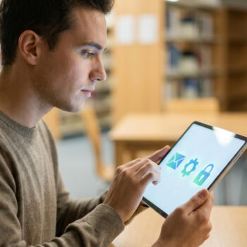 Étudiant concentré utilisant une tablette avec des icônes d'e-mail, de réglages et de cadenas dans une bibliothèque.