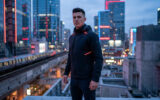 Un homme à la coiffure soignée portant une veste tactique avec des détails lumineux dans une ville futuriste.