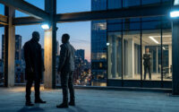 Deux hommes font face à un homme en costume derrière une vitre sur un toit terrasse au crépuscule.