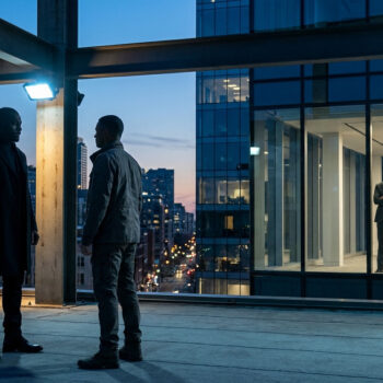 Deux hommes font face à un homme en costume derrière une vitre sur un toit terrasse au crépuscule.