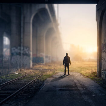 Une silhouette seule avec un sac à dos se tient sur une voie ferrée sous un pont recouvert de graffitis au coucher du soleil.