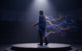 Un homme debout sur une scène circulaire, enveloppé par des traînées de fumée colorée sous un projecteur.