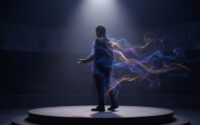 Un homme debout sur une scène circulaire, enveloppé par des traînées de fumée colorée sous un projecteur.