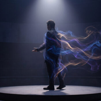 Un homme debout sur une scène circulaire, enveloppé par des traînées de fumée colorée sous un projecteur.