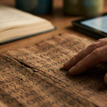 Main touchant un papyrus ancien avec un livre et une tablette en arrière-plan.