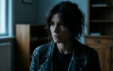 Portrait d'une femme aux cheveux bruns ébouriffés portant une veste en cuir, regardant au loin avec une expression sombre.