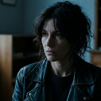 Portrait d'une femme aux cheveux bruns ébouriffés portant une veste en cuir, regardant au loin avec une expression sombre.