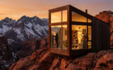 Une cabine moderne en verre sur une falaise face à des montagnes enneigées sous un coucher de soleil doré.