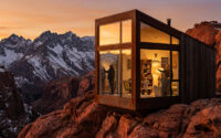 Une cabine moderne en verre sur une falaise face à des montagnes enneigées sous un coucher de soleil doré.