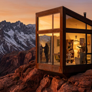 Une cabine moderne en verre sur une falaise face à des montagnes enneigées sous un coucher de soleil doré.