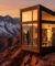 Une cabine moderne en verre sur une falaise face à des montagnes enneigées sous un coucher de soleil doré.