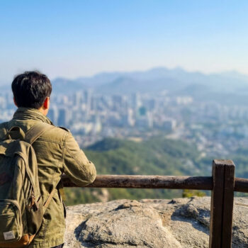 Un jeune homme avec un sac à dos regarde une ville floue depuis un sommet montagneux.