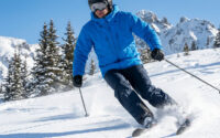 Skieur en veste bleue et pantalon noir dévalant une piste enneigée en montagne.