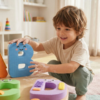 Un jeune enfant souriant joue avec des lettres colorées en mousse sur une petite table en bois dans une chambre.