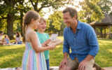 Deux enfants souriants offrent un cadeau emballé et une carte dessinée à leur père dans un parc ensoleillé.