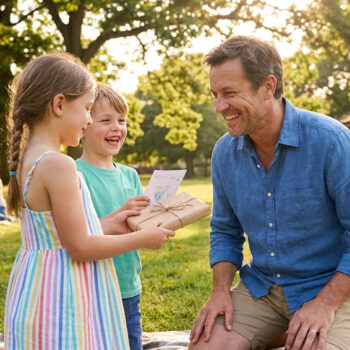 Deux enfants souriants offrent un cadeau emballé et une carte dessinée à leur père dans un parc ensoleillé.