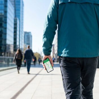 Un homme marche dans une rue moderne en consultant l'application Mappy sur son smartphone pour s'orienter.