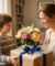 Un jeune garçon souriant offre un bouquet de fleurs et un cadeau emballé à sa mère dans un salon lumineux.
